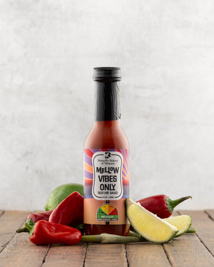 Bottle of Butterfly Bakery of Vermont Mellow Vibes Only Hot Sauce on a wood surface alongside red jalapenos, garlic scapes and lime wedges