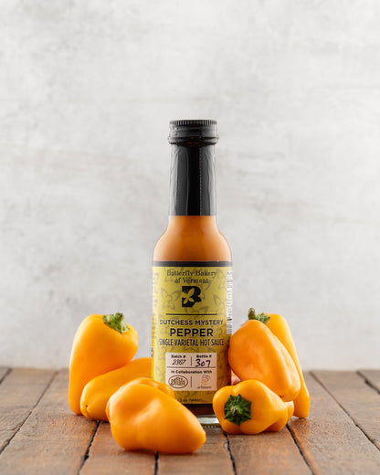 A bottle of Butterfly Bakery of Vermont Dutchess Mystery Pepper Hot Sauce on a wood surface, surrounded by yellow hot peppers