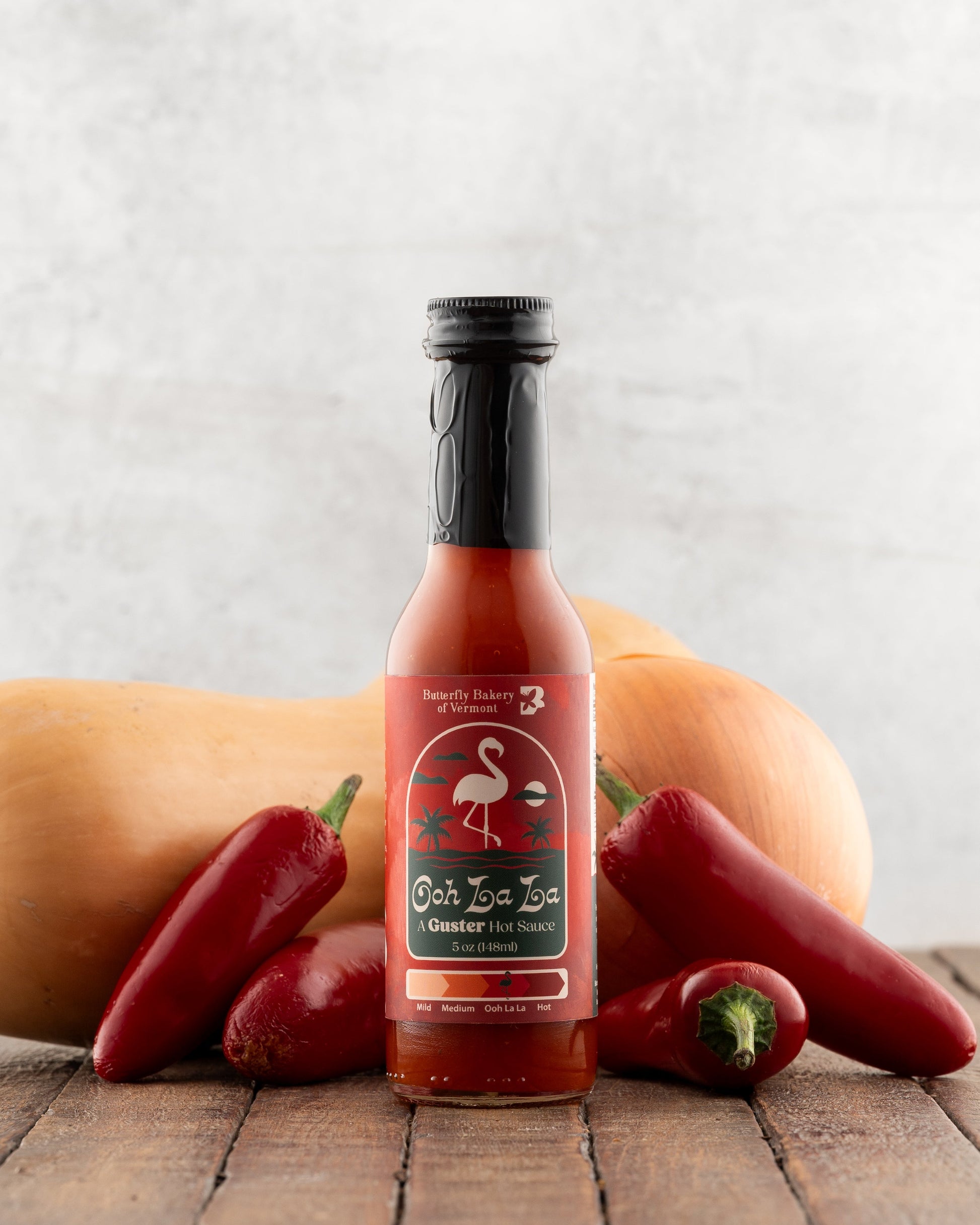 Bottle of Butterfly Bakery of Vermont Ooh La La Hot Sauce on a wood surface with red jalapenos and a butternut squash in the background.
