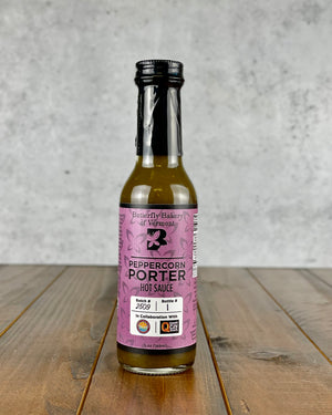 A bottle of Peppercorn Porter Hot Sauce by Butterfly Hakes from Vermont, featuring a purple label and black cap, set on a rustic wooden surface against a textured gray concrete wall.