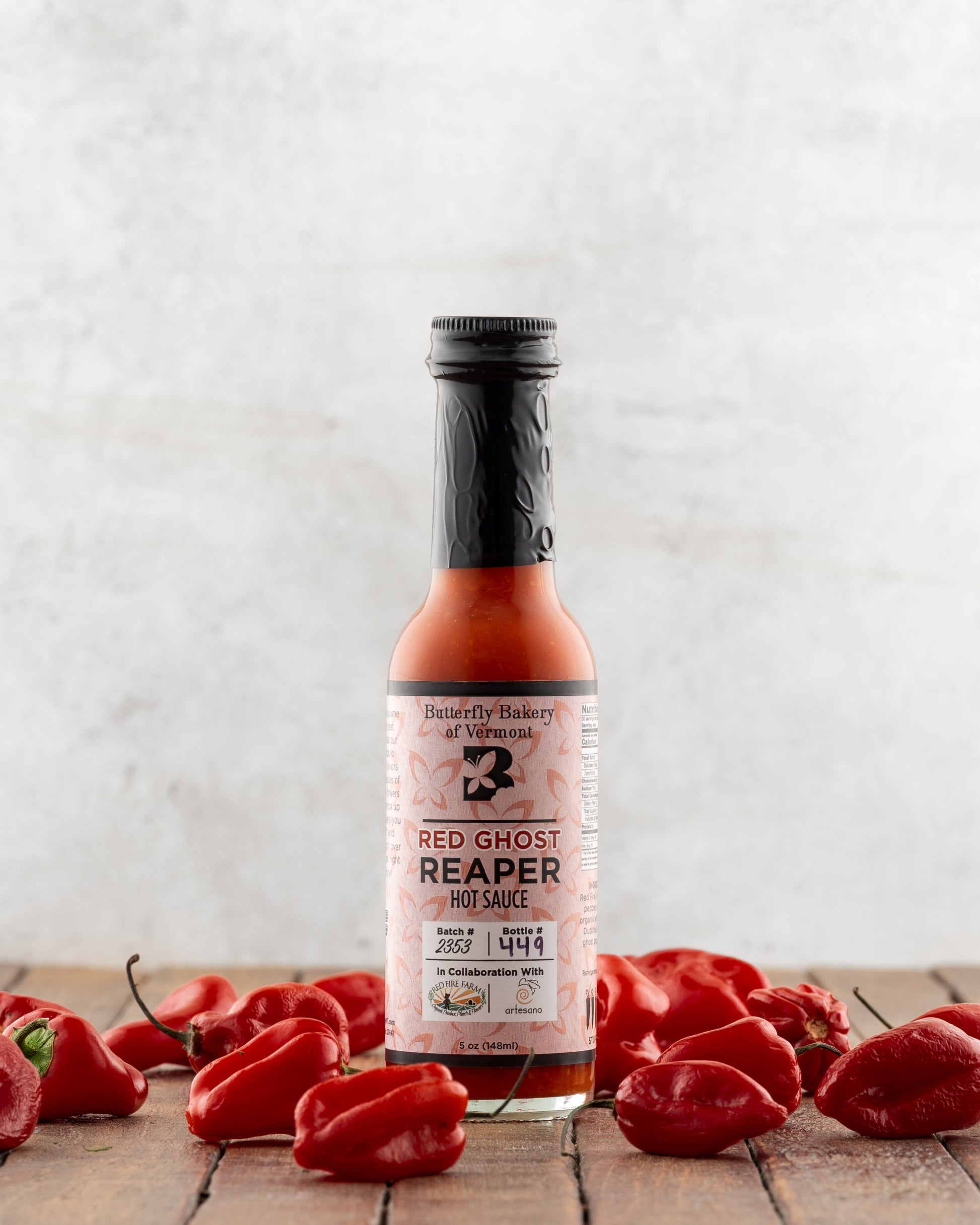 Bottle of Butterfly Bakery of Vermont Red Ghost Reaper Hot Sauce surrounded by red ghost peppers on a wooden surface