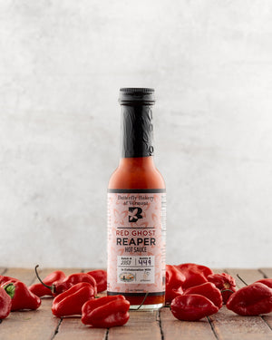Bottle of Butterfly Bakery of Vermont Red Ghost Reaper Hot Sauce surrounded by red ghost peppers on a wooden surface