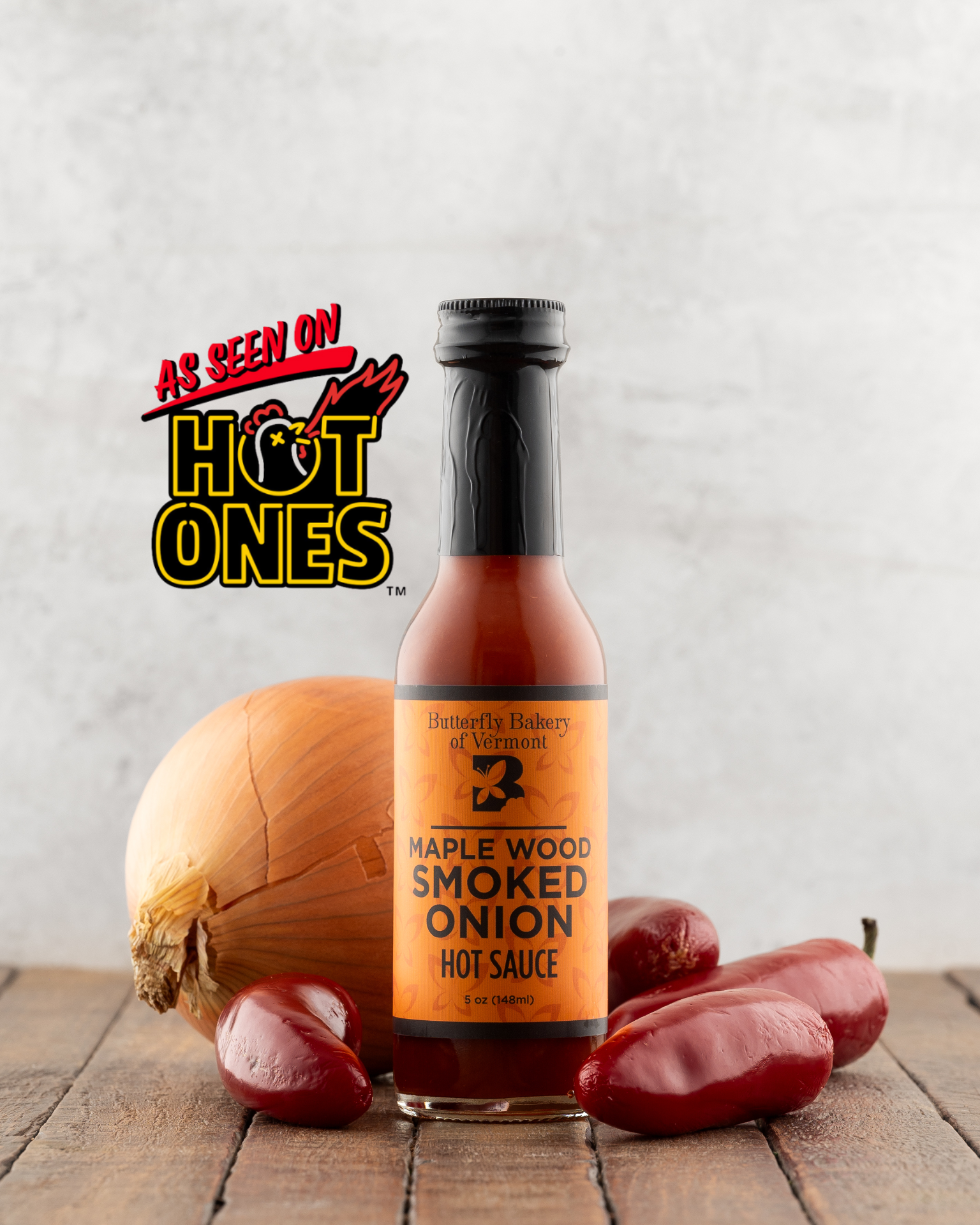 Bottle of Butterfly Bakery of Vermont Maple Wood Smoked Onion Hot Sauce on a wood surface with an onion and jalapeno peppers, with the 'As Seen On Hot Ones' logo