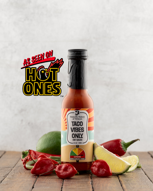Bottle of Butterfly Bakery of Vermont Taco Vibes Only Hot Sauce aside lime, hot peppers and garlic scapes on a wood surface and the 'As Seen on Hot Ones' logo.
