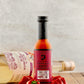 Bottle of Elderflower Ghost Craft Spirit hot sauce on a wooden surface with peppers and a bottle of rum in the background
