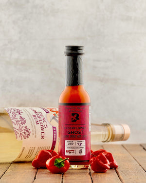 Bottle of Elderflower Ghost Craft Spirit hot sauce on a wooden surface with peppers and a bottle of rum in the background