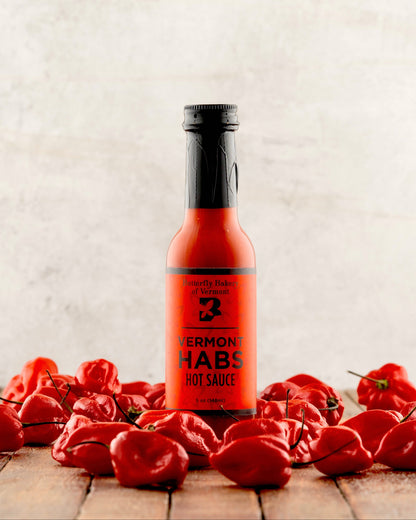 Bottle of Vermont Habs hot sauce surrounded by red peppers on a wooden surface.