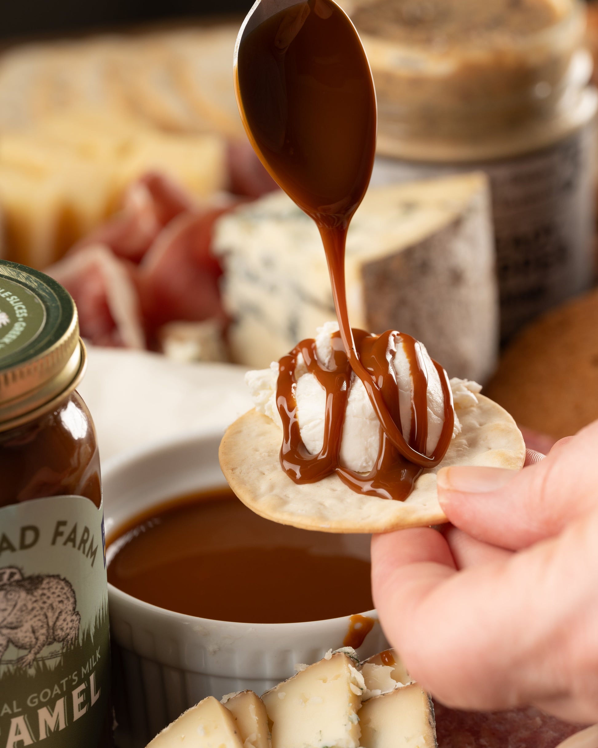 Person drizzling caramel onto a cracker topped with goat cheese with various crackers and cheese in the background.