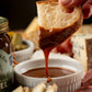 Dipping bread into a bowl of caramel sauce with a jar of caramel and other snacks in the background.