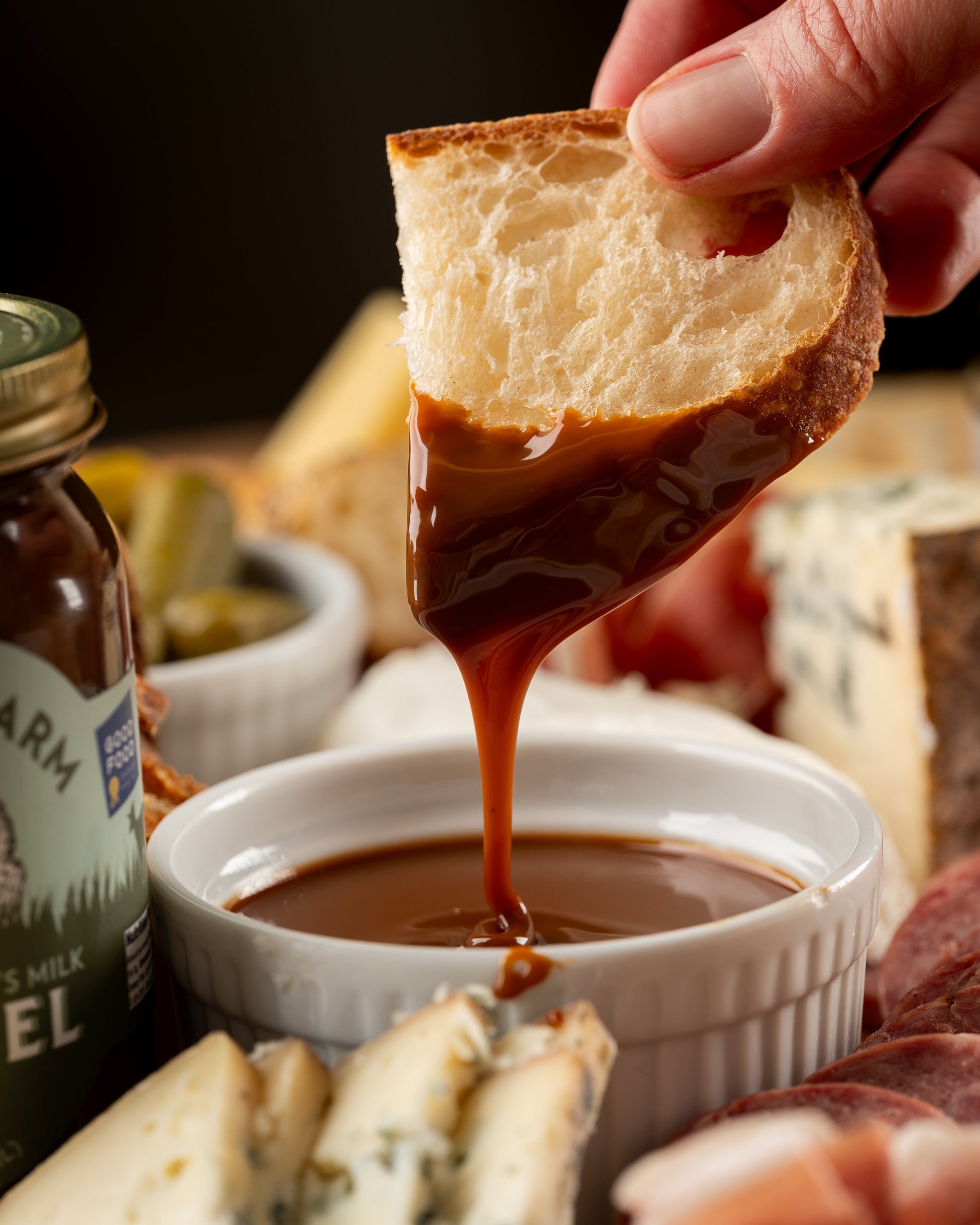 Dipping bread into a bowl of caramel sauce with a jar of caramel and other snacks in the background.