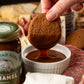 Person dipping a cookie into caramel sauce with a charcuterie board in the background
