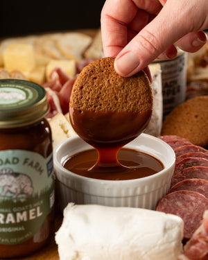 Person dipping a cookie into caramel sauce with a charcuterie board in the background
