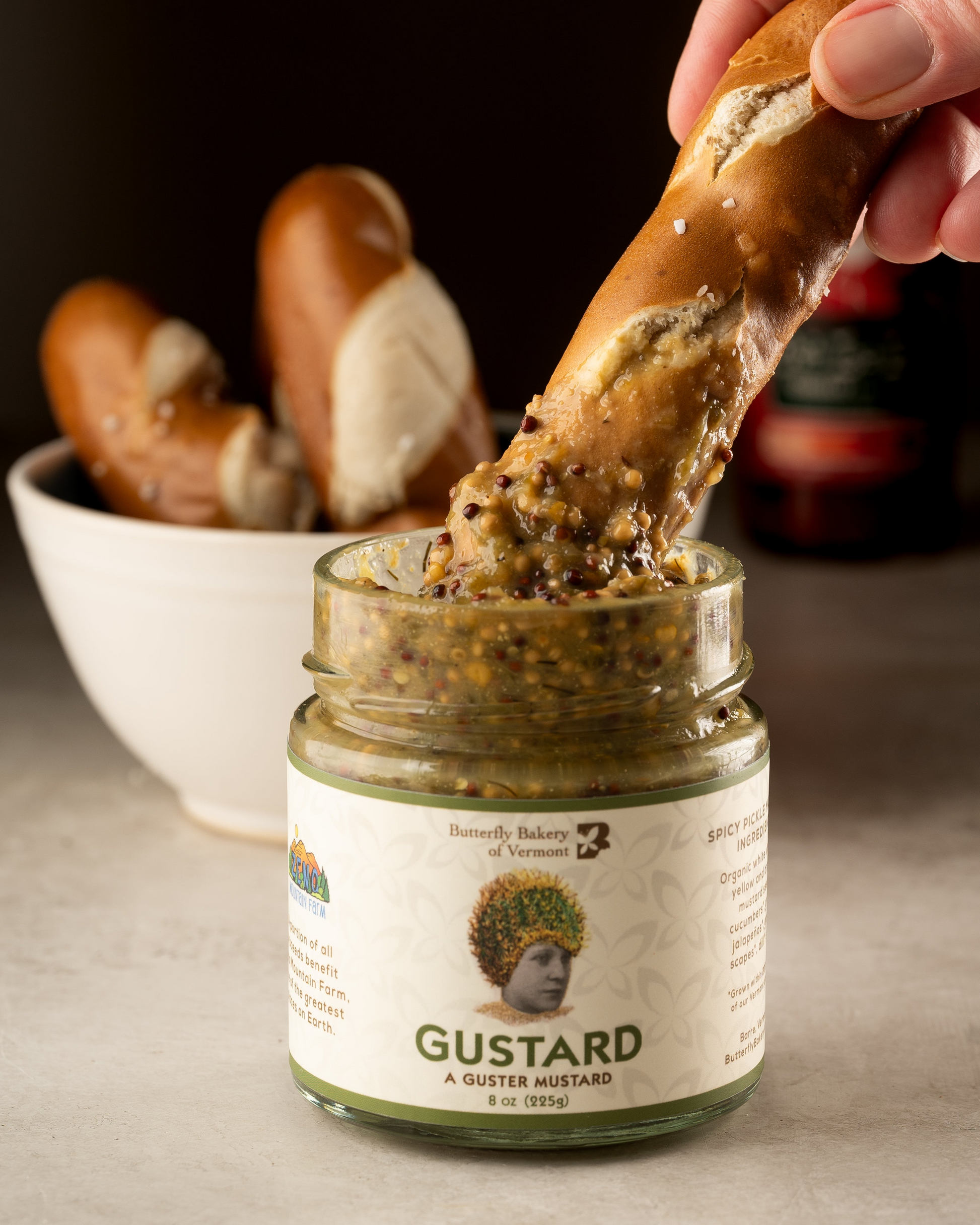 A hand dipping a soft pretzel stick into a jar of Gustard Mustard from Butterfly Bakery of Vermont