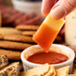 Hand dipping a piece of cheese into a small bowl of hot sauce with crackers and cheese in the background.