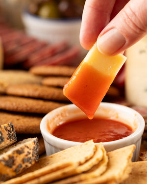 Hand dipping a piece of cheese into a small bowl of hot sauce with crackers and cheese in the background.