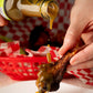 Person pouring hot sauce onto a chicken wing on a white plate with a red checkered background.