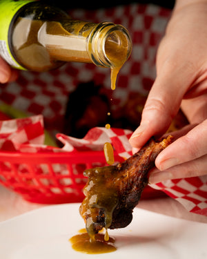 Person pouring hot sauce onto a chicken wing on a white plate with a red checkered background.