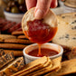 Hand dipping a cracker and pepperoni into a small bowl of hot sauce with various crackers and cheese in the background.