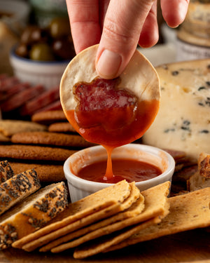 Hand dipping a cracker and pepperoni into a small bowl of hot sauce with various crackers and cheese in the background.