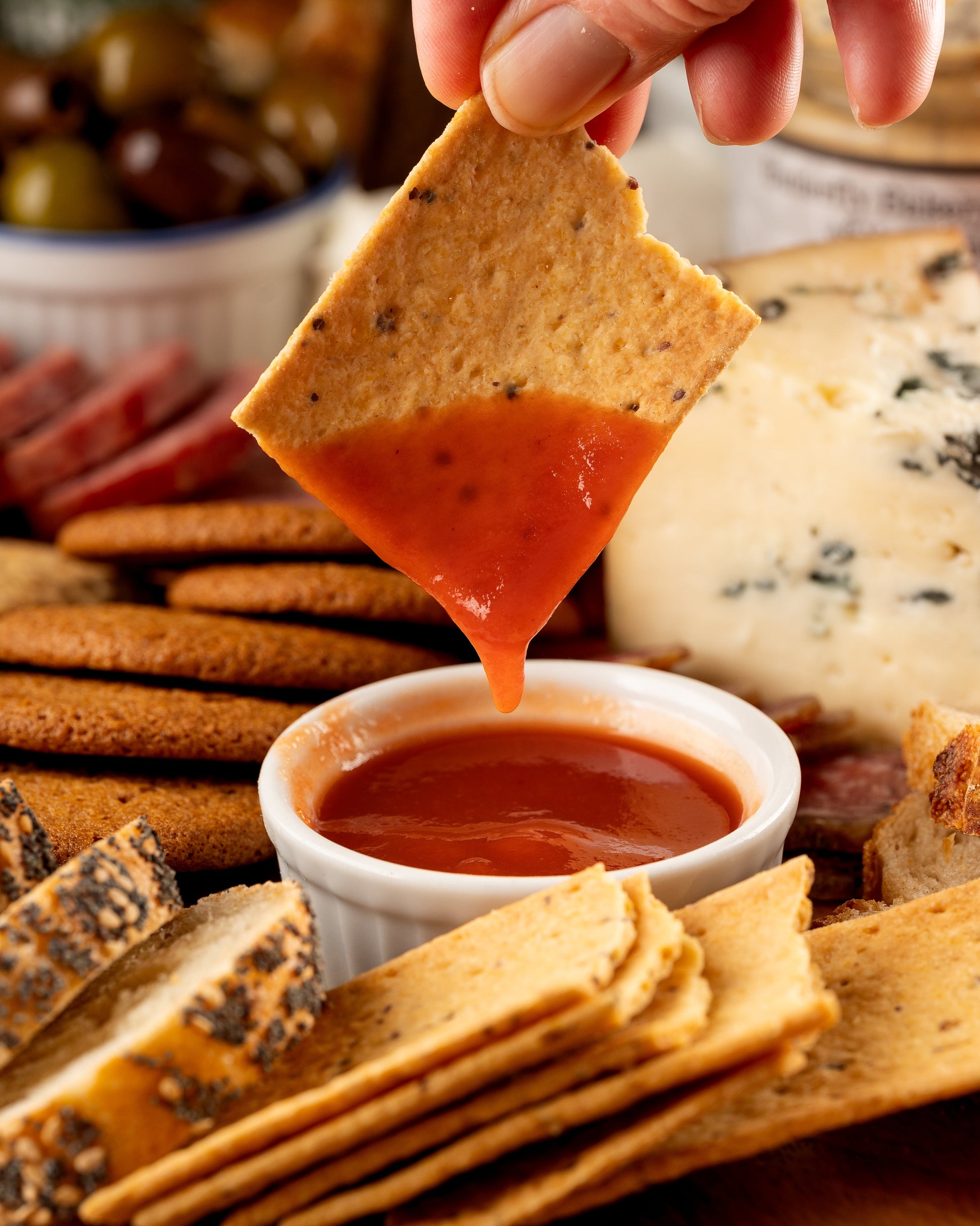 Hand dipping a cracker into a small bowl of hot sauce with a platter of crackers and cheese in the background.