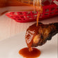 Hot sauce being poured onto a chicken wing with a basket lined with red and white checkered paper in the background