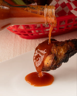 Hot sauce being poured onto a chicken wing with a basket lined with red and white checkered paper in the background