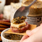 Spoon holding a small appetizer with cheese, salami, and pickles over a ramekin of the mustard being spooned onto the appetizer.