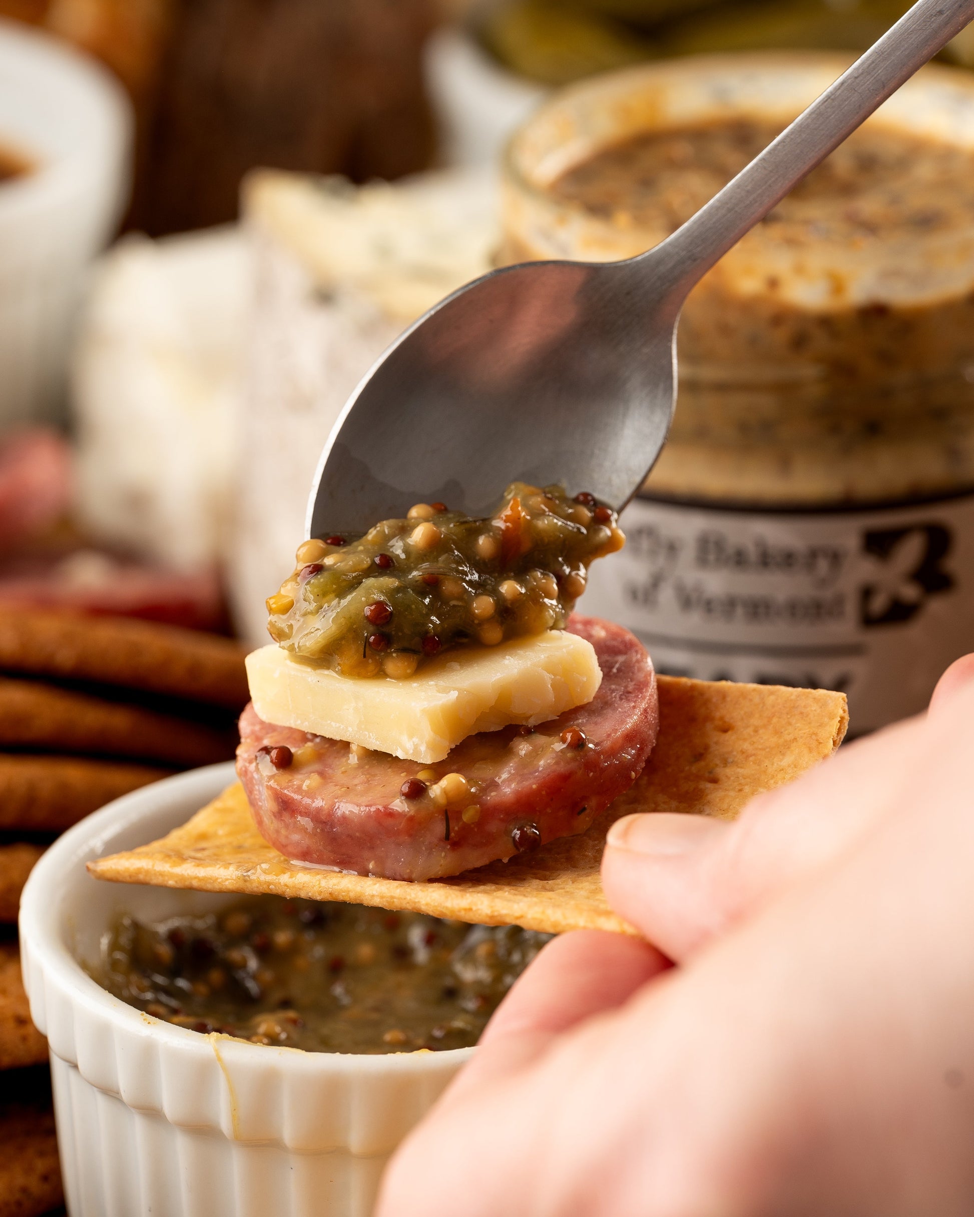 Spoon holding a small appetizer with cheese, salami, and pickles over a ramekin of the mustard being spooned onto the appetizer.