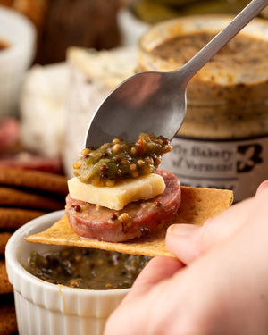 Spoon holding a small appetizer with cheese, salami, and pickles over a ramekin of the mustard being spooned onto the appetizer.