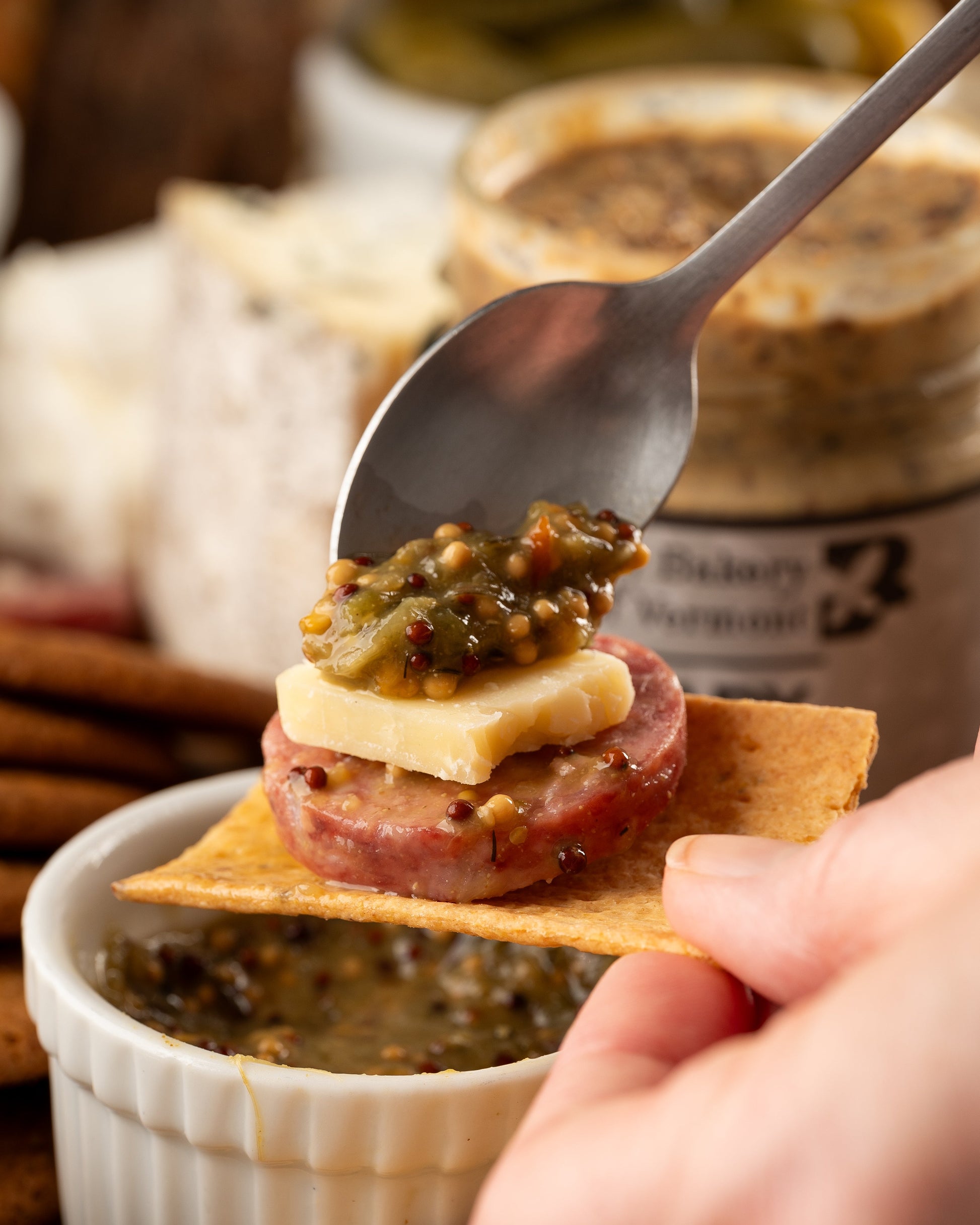Hand holding a cracker with salami, cheese, and mustard being spooned on top.