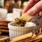 Piece of hard cheese being dipped into a small bowl of Butterfly Bakery of Vermont Heady Topper mustard surrounded by crackers, bread and cheese