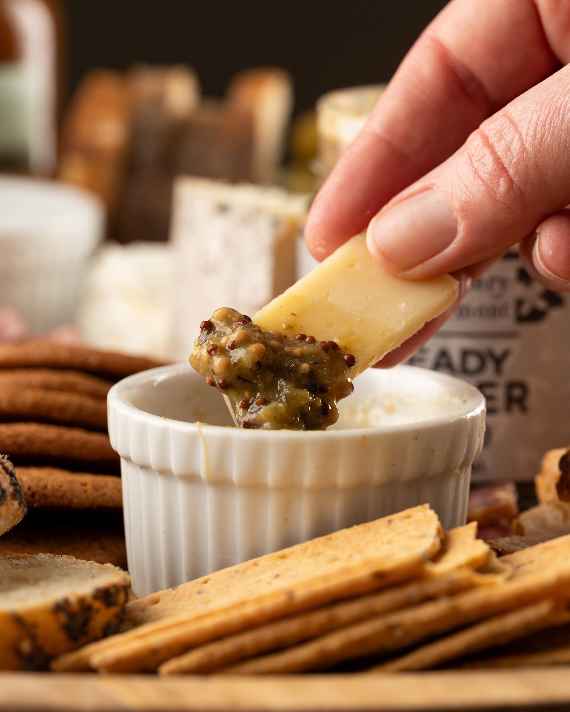 Piece of hard cheese being dipped into a small bowl of Butterfly Bakery of Vermont Heady Topper mustard surrounded by crackers, bread and cheese