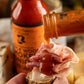 Hot sauce being poured onto a slice of bread with cheese and ham, with a bottle of hot sauce in the background.