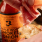 Person adding prosciutto to a piece of bread with a bottle of 'Butterfly Bakery of Vermont Maple Wood Smoked Onion' hot sauce in the background.