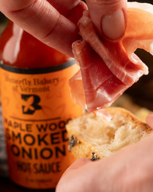 Person adding prosciutto to a piece of bread with a bottle of 'Butterfly Bakery of Vermont Maple Wood Smoked Onion' hot sauce in the background.