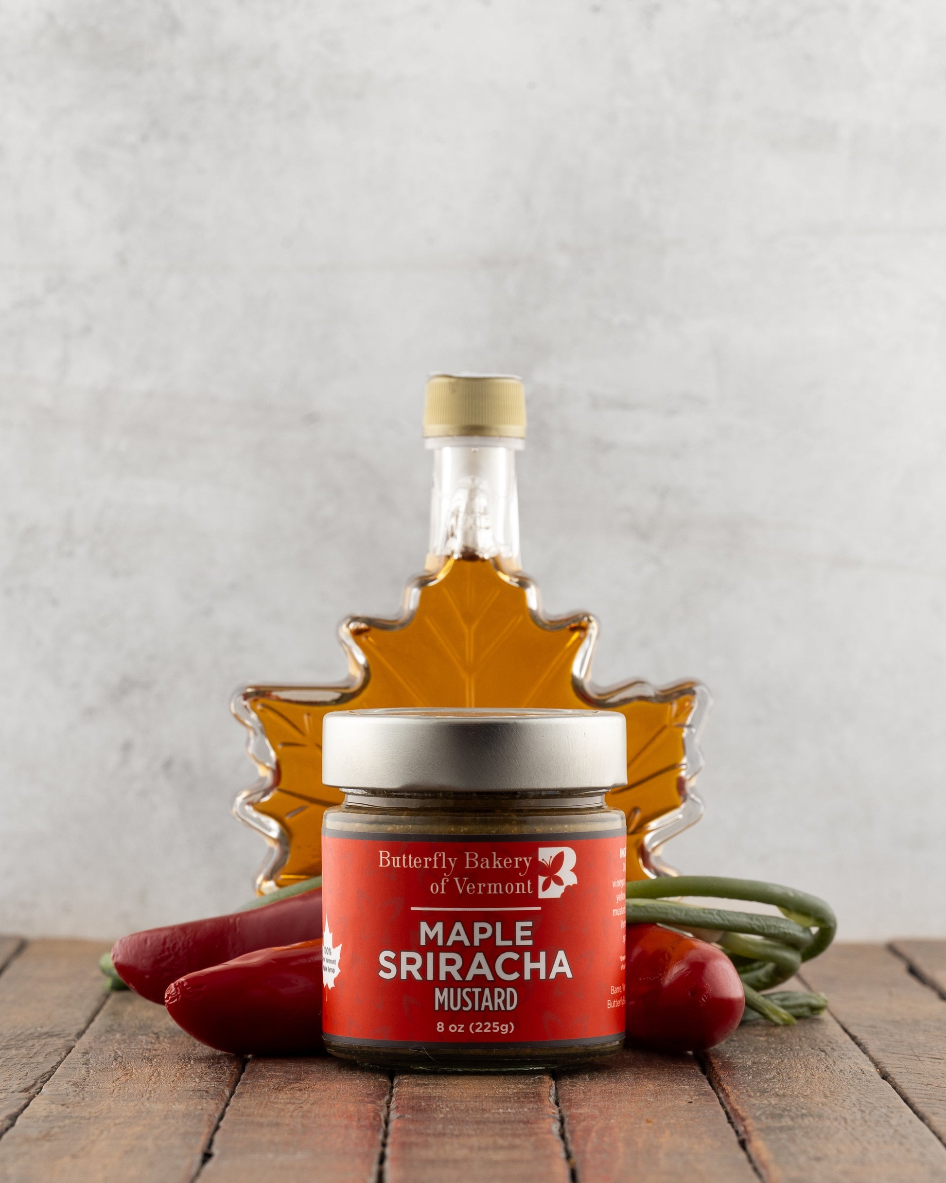 Butterfly Bakery of Vermont Maple Sriracha Mustard on a wood surface with red peppers and a bottle of maple syrup