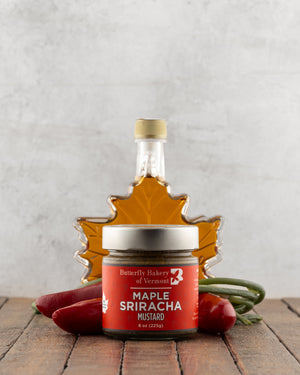 Butterfly Bakery of Vermont Maple Sriracha Mustard on a wood surface with red peppers and a bottle of maple syrup
