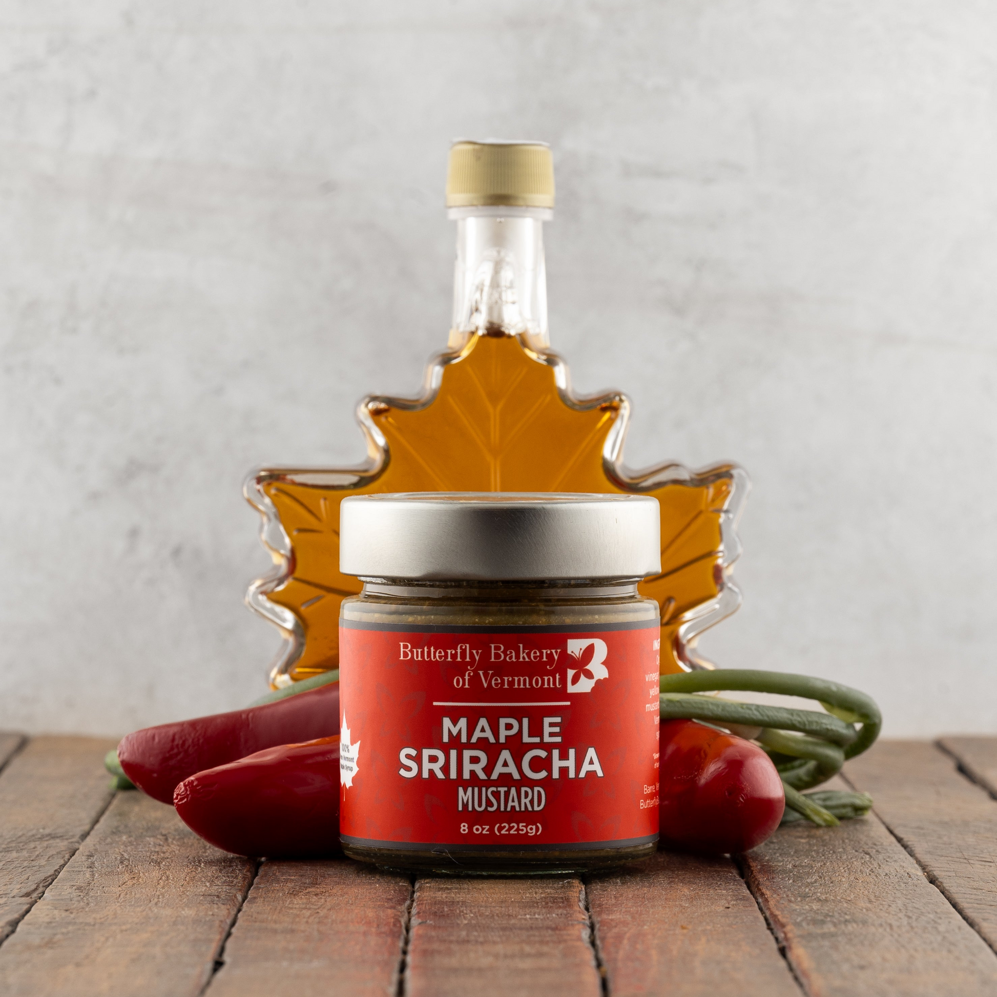 Jar of Maple Sriracha Mustard with a bottle of maple syrup and vegetables on a wooden surface.