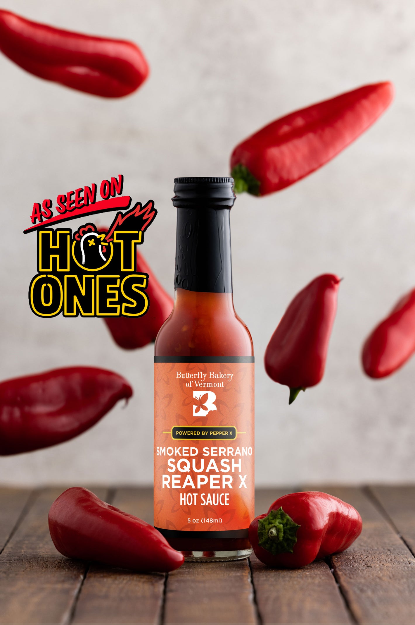 Bottle of Butterfly Bakery of Vermont's Smoked Serrano Squash Reaper X hot sauce surrounded by red hot peppers on a wooden surface with the 'As Seen On Hot Ones' logo.