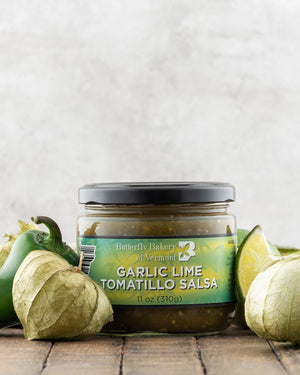 Butterfly Bakery of Vermont Garlic Lime Tomatillo Salsa on a wood surface with green peppers, tomatillos and a lime wedge