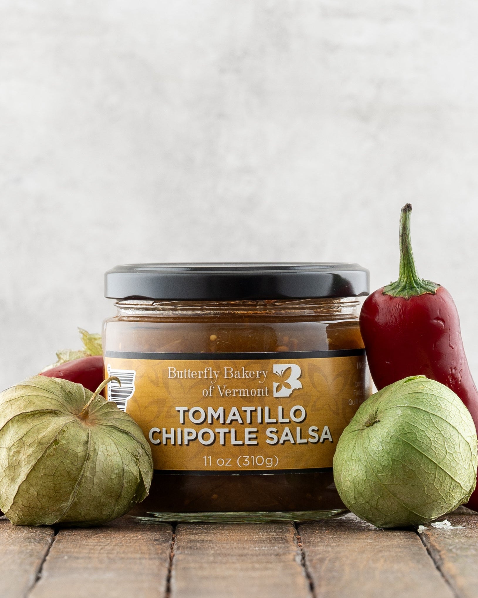 Butterfly Bakery of Vermont Tomatillo Chipotle Salsa on a wood surface with red peppers and tomatillos