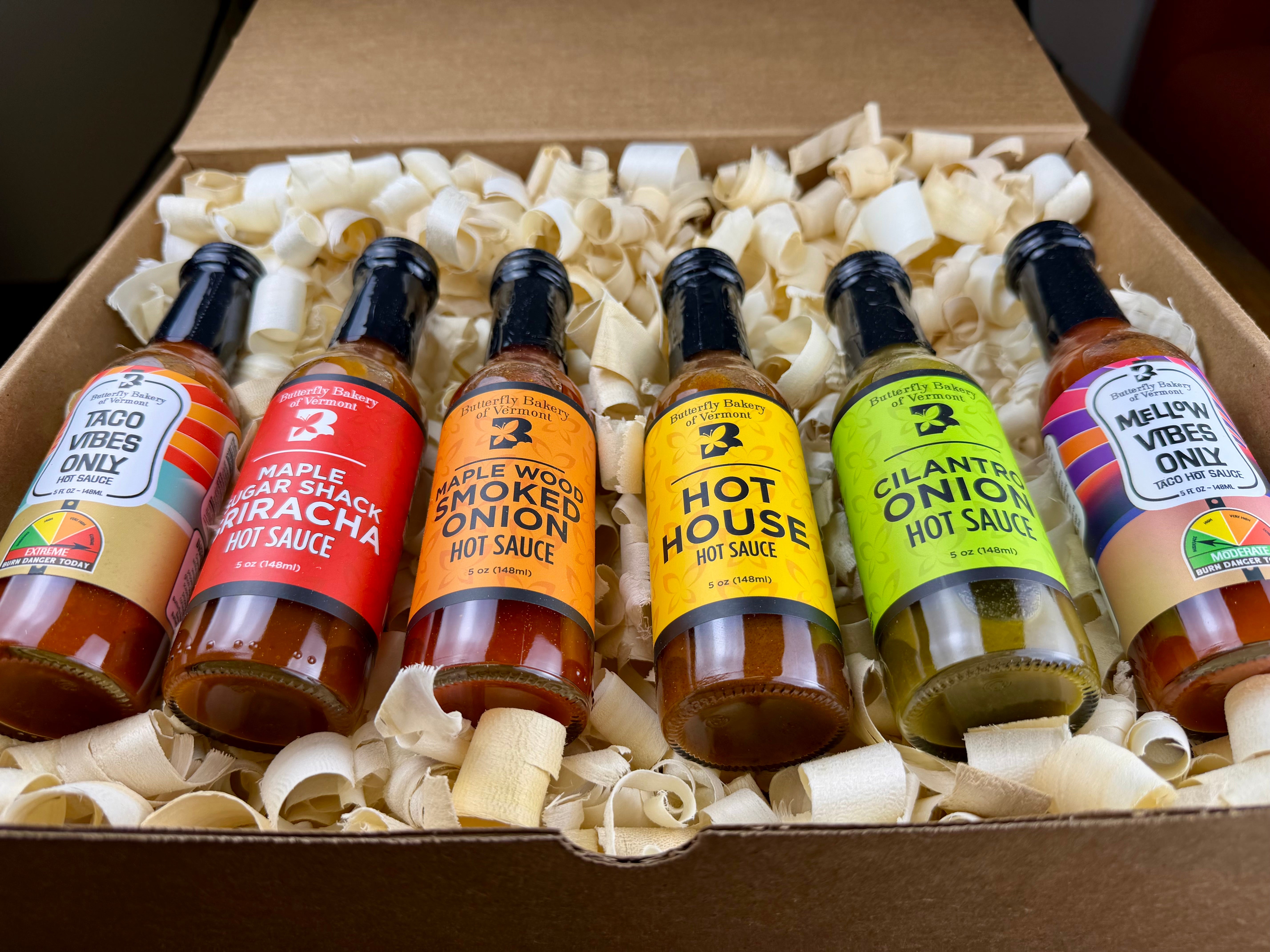 Six bottles of Butterfly Bakery of Vermont hot sauce in a box with decorative wood shavings
