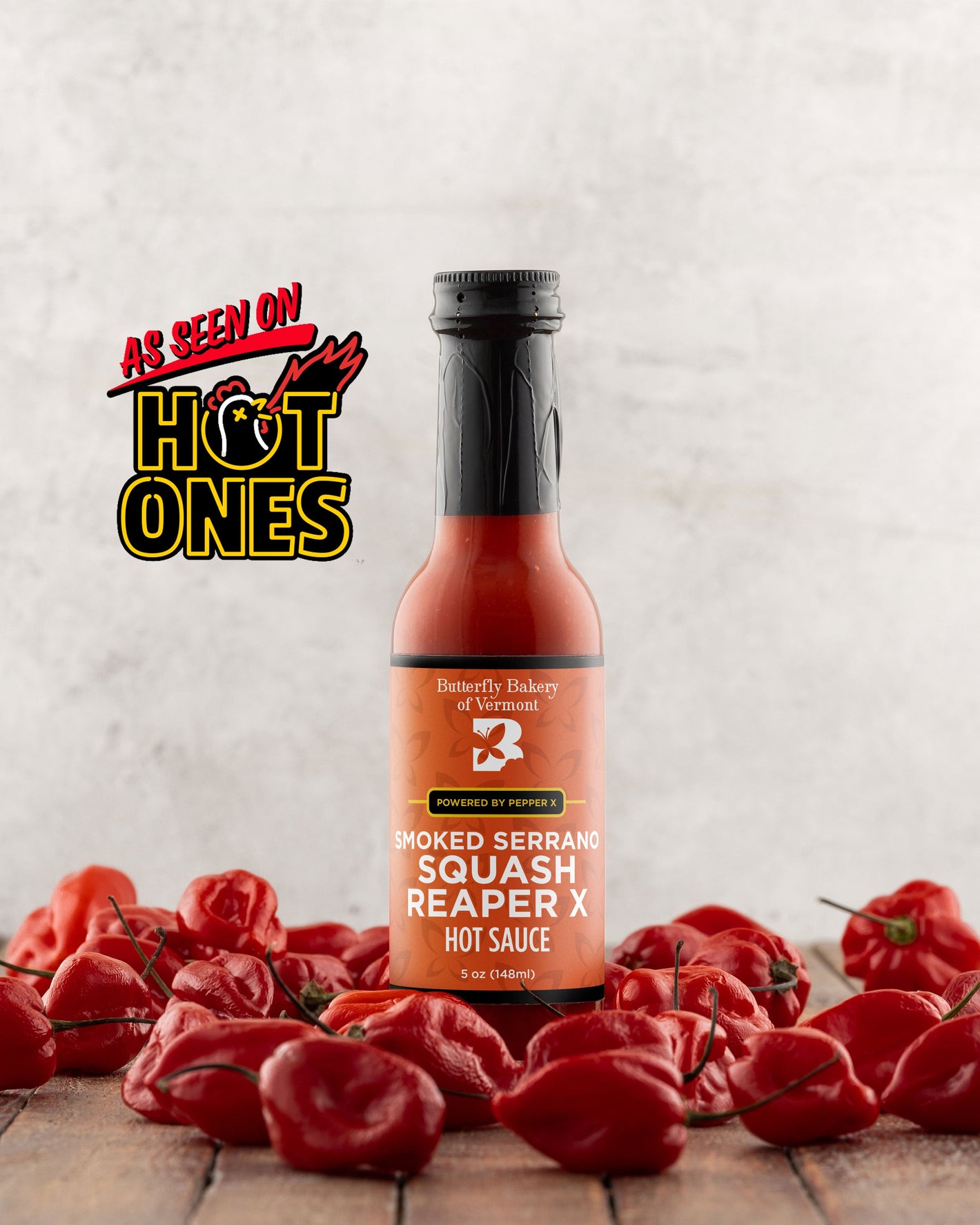 Bottle of Butterfly Bakery of Vermont's Smoked Serrano Squash Reaper X hot sauce surrounded by red hot peppers on a wooden surface with the 'As Seen On Hot Ones' logo.