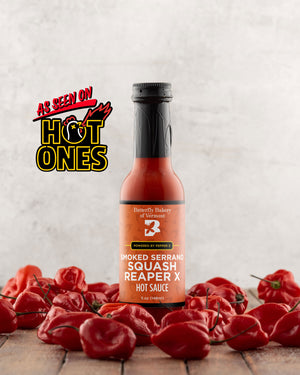 Bottle of Butterfly Bakery of Vermont's Smoked Serrano Squash Reaper X hot sauce surrounded by red hot peppers on a wooden surface with the 'As Seen On Hot Ones' logo.