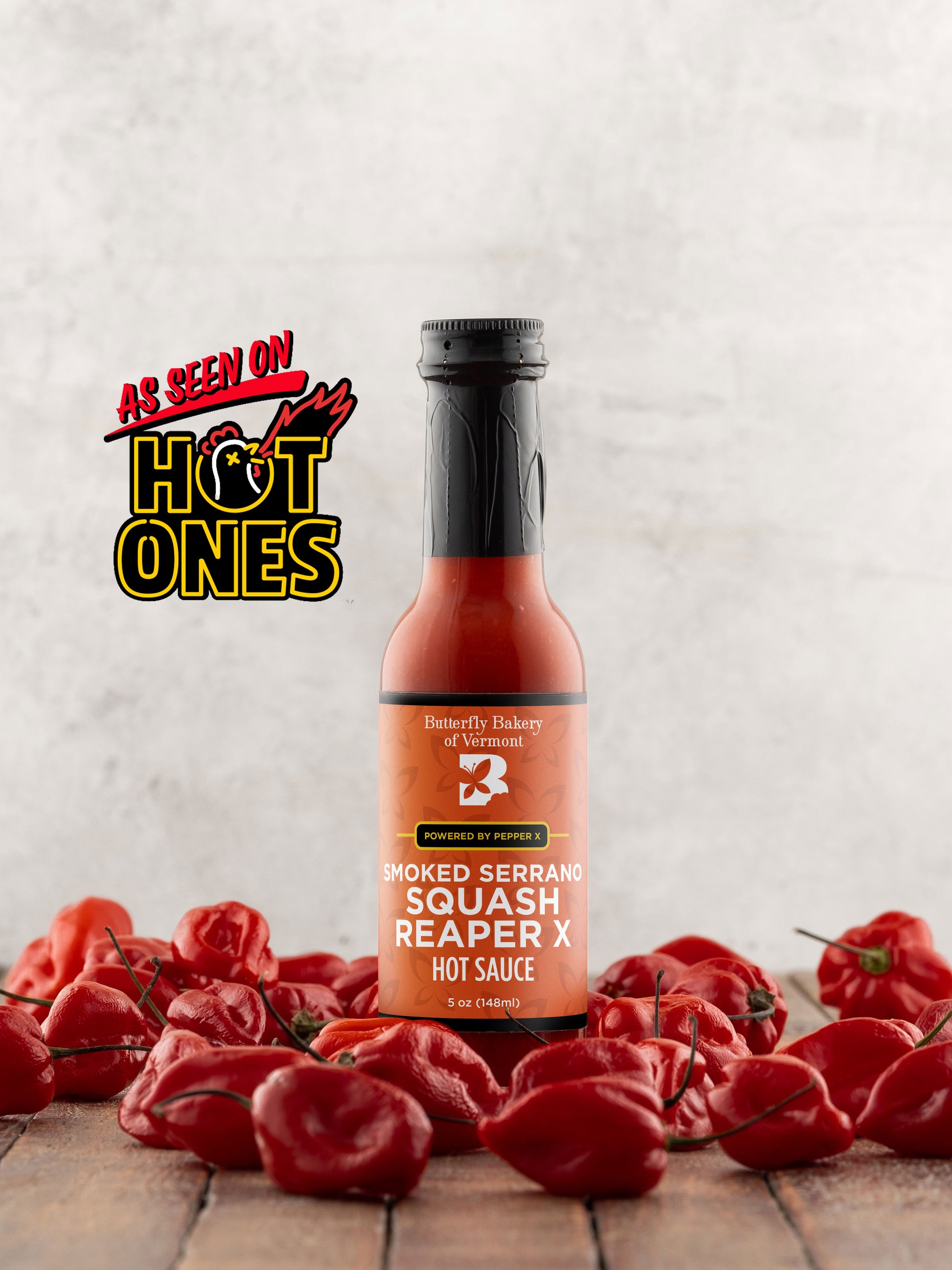 Bottle of Butterfly Bakery of Vermont Smoked Serrano Squash Reaper X Hot Sauce surrounded by red hot peppers on a wooden surface with the 'As Seen On Hot Ones' logo