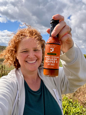 Claire Georges holding a bottle of Smoked Serrano Squash Reaper X hot sauce in a  farm field