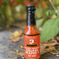 A bottle of Smoked Serrano Squash Reaper X hot sauce sitting on the ground in front of a hot pepper plant