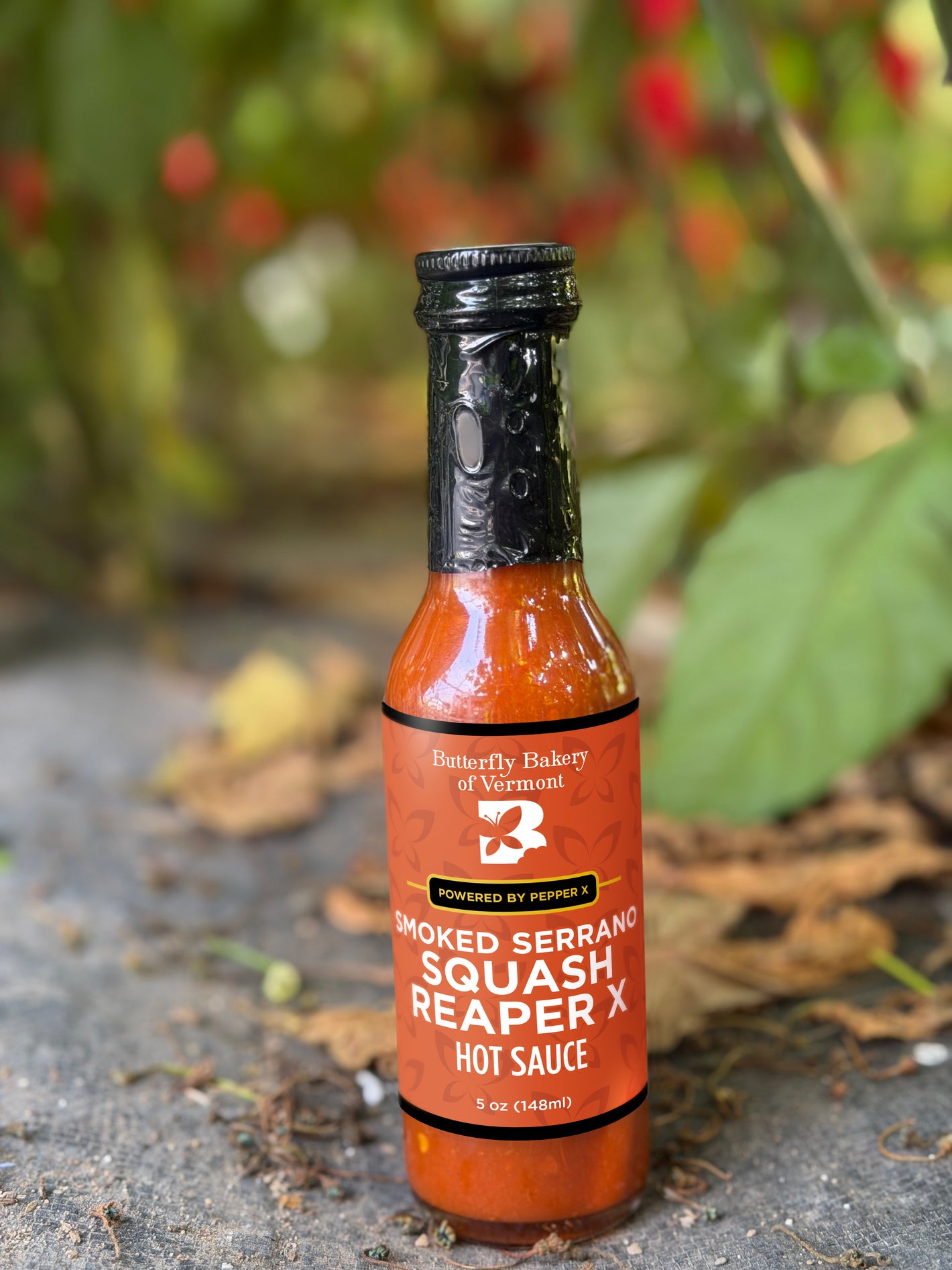A bottle of Smoked Serrano Squash Reaper X hot sauce sitting on the ground in front of a hot pepper plant