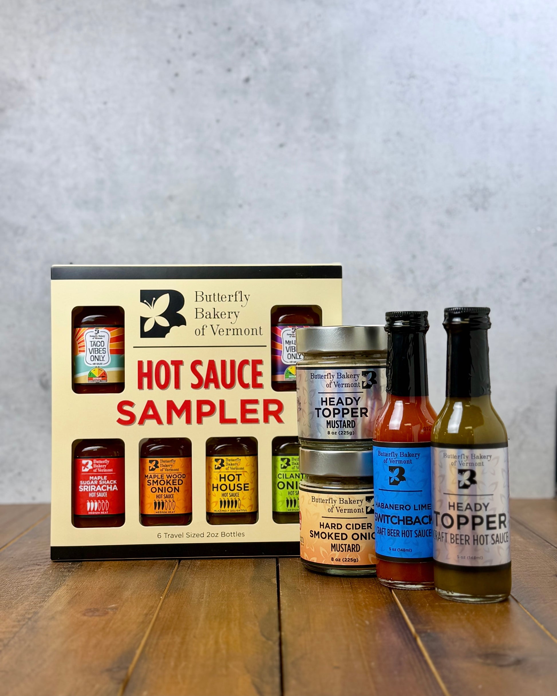 A Butterfly Bakery of Vermont hot sauce sampler box, along with 2 other bottles of hot sauce and 2 jars of mustard on a wood surface and gray background