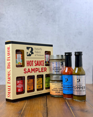 A Butterfly Bakery of Vermont hot sauce sampler box, along with 2 other bottles of hot sauce and 2 jars of mustard on a wood surface and gray background